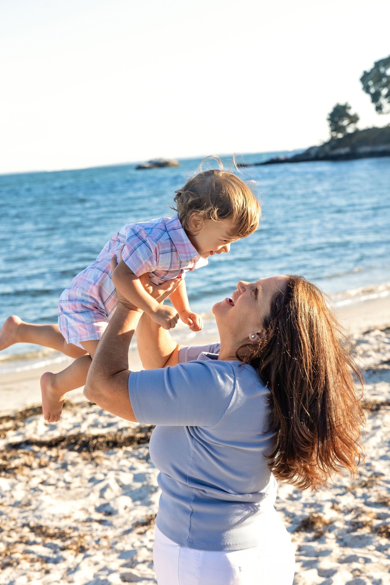 A parent and child share a joyful moment playing together on a sunny beach with ocean waves in the background.