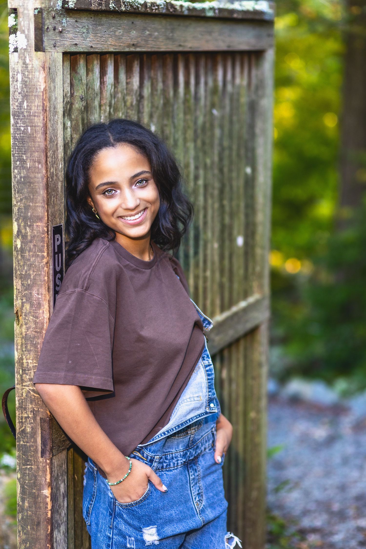 A casual outdoor photo session against a wooden fence featuring denim overalls and a brown t-shirt on a sunny day.