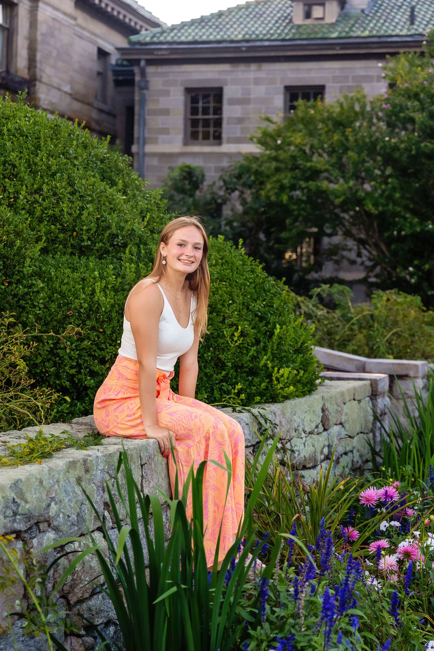 Someone sits on a stone wall in a garden with colorful flowers and greenery near a historic stone building.