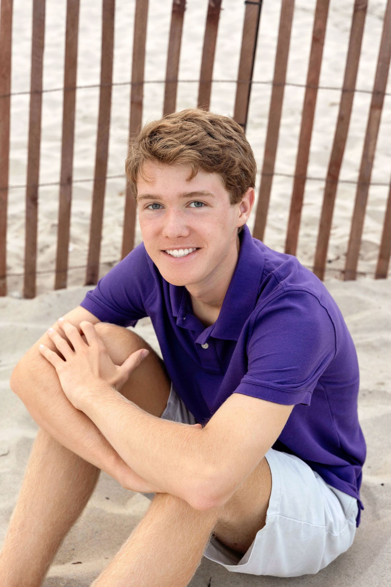 A person in a purple polo shirt and white shorts lies casually on a sandy beach near a wooden fence.
