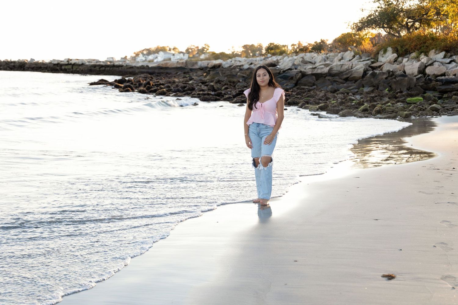 Person walking along a scenic rocky beach shoreline with waves and rocks at sunset.