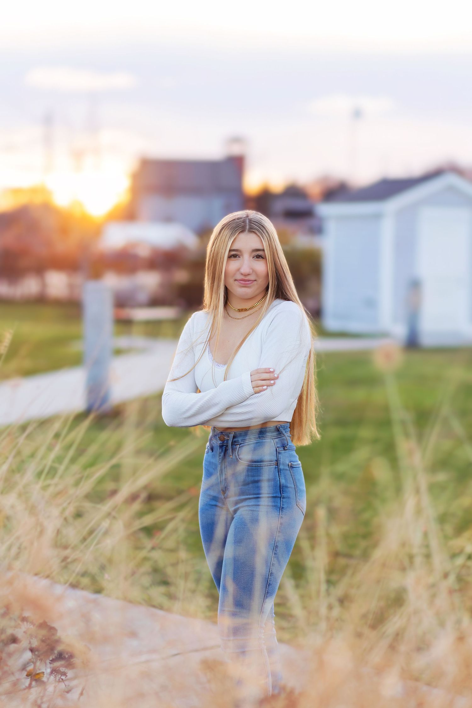 A person in a white sweater and jeans poses in a grassy field during golden hour sunset.
