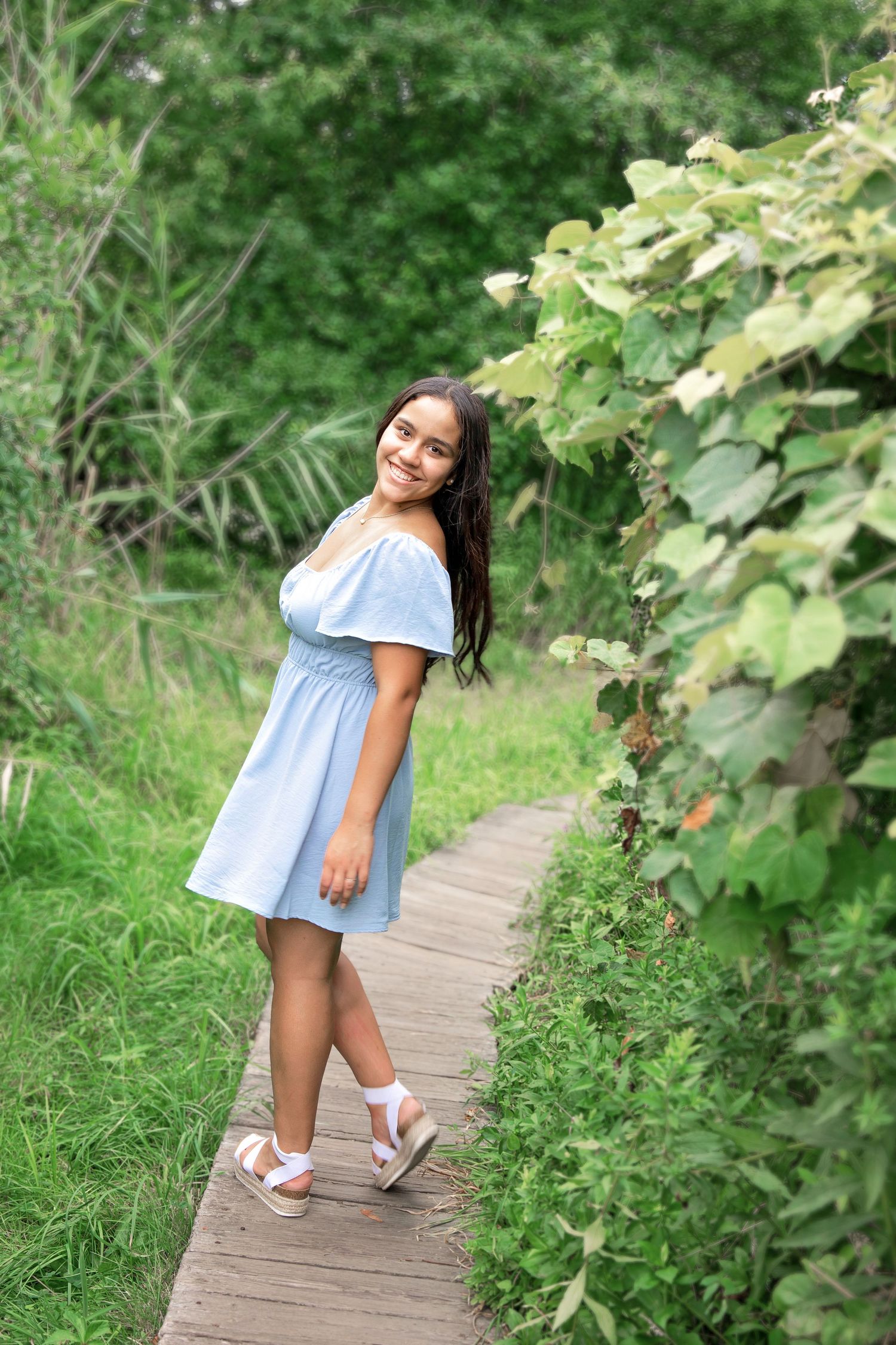 A person in a light blue dress walks along a wooden boardwalk path surrounded by lush green foliage on a summer day.