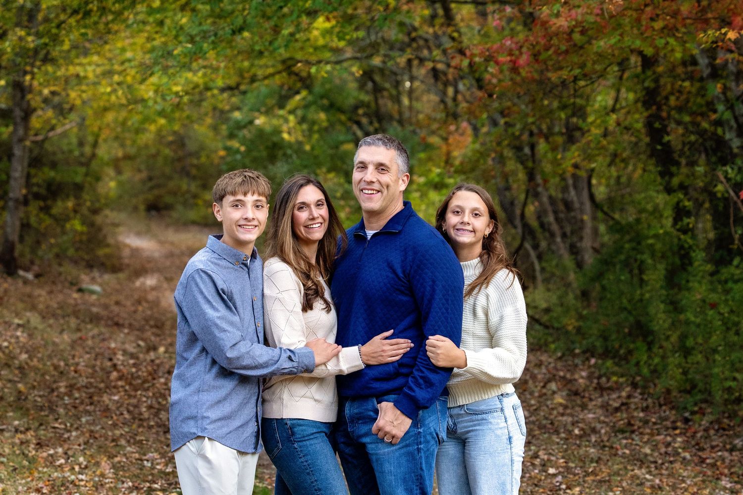 A family wearing coordinated blue and white outfits poses together outdoors among autumn trees and foliage.