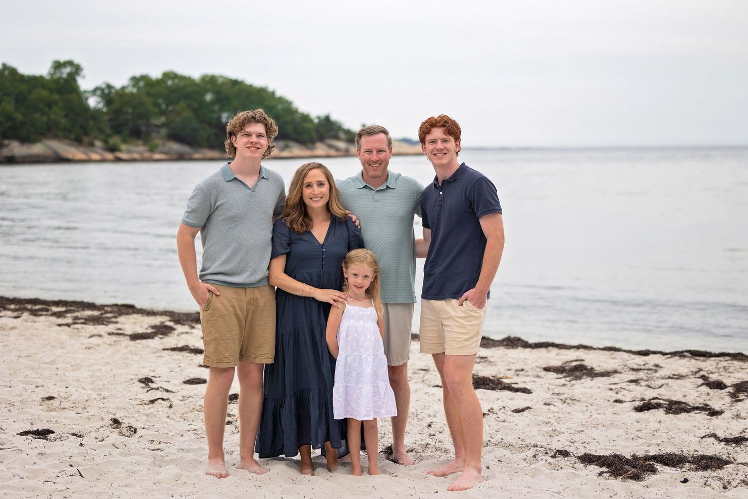 A group of people wearing casual clothing pose together on a sandy beach at sunset.