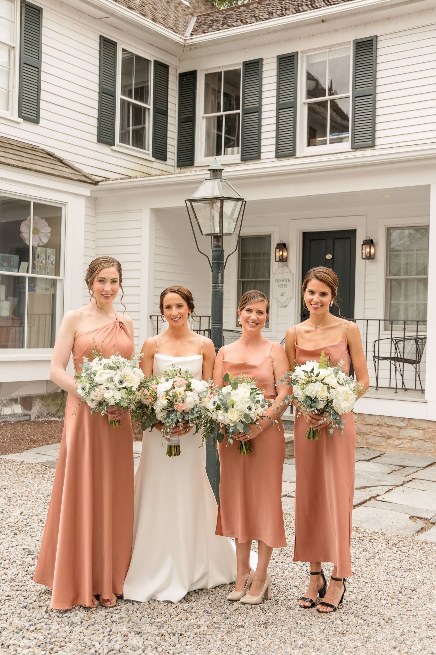 Bridesmaids in dusty rose colored dresses stand in line holding white floral bouquets outside a white colonial style house.