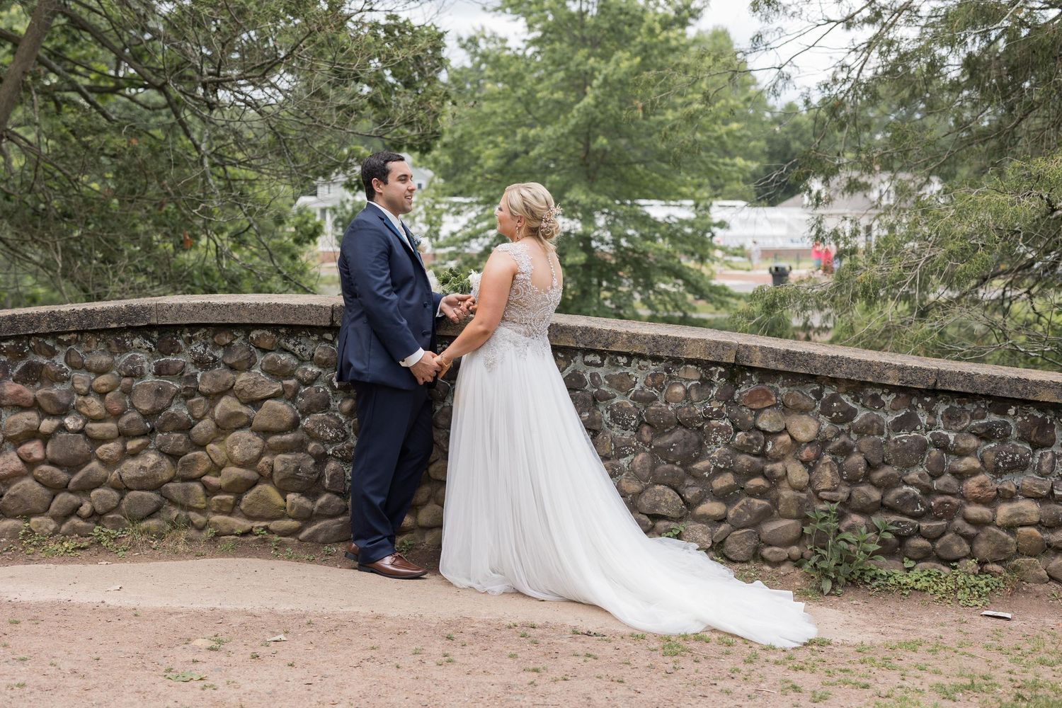 A couple in formal wedding attire stands together on a rustic stone wall path surrounded by lush greenery.