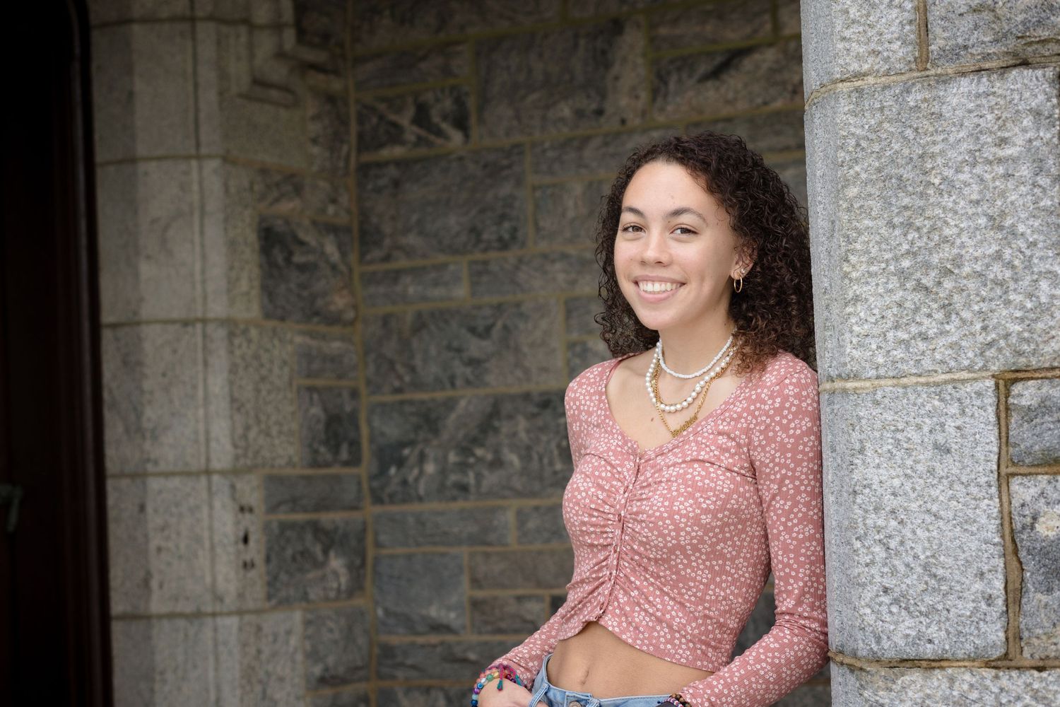 Woman in pink floral dress smiles while sitting on stone steps near a stone wall.
