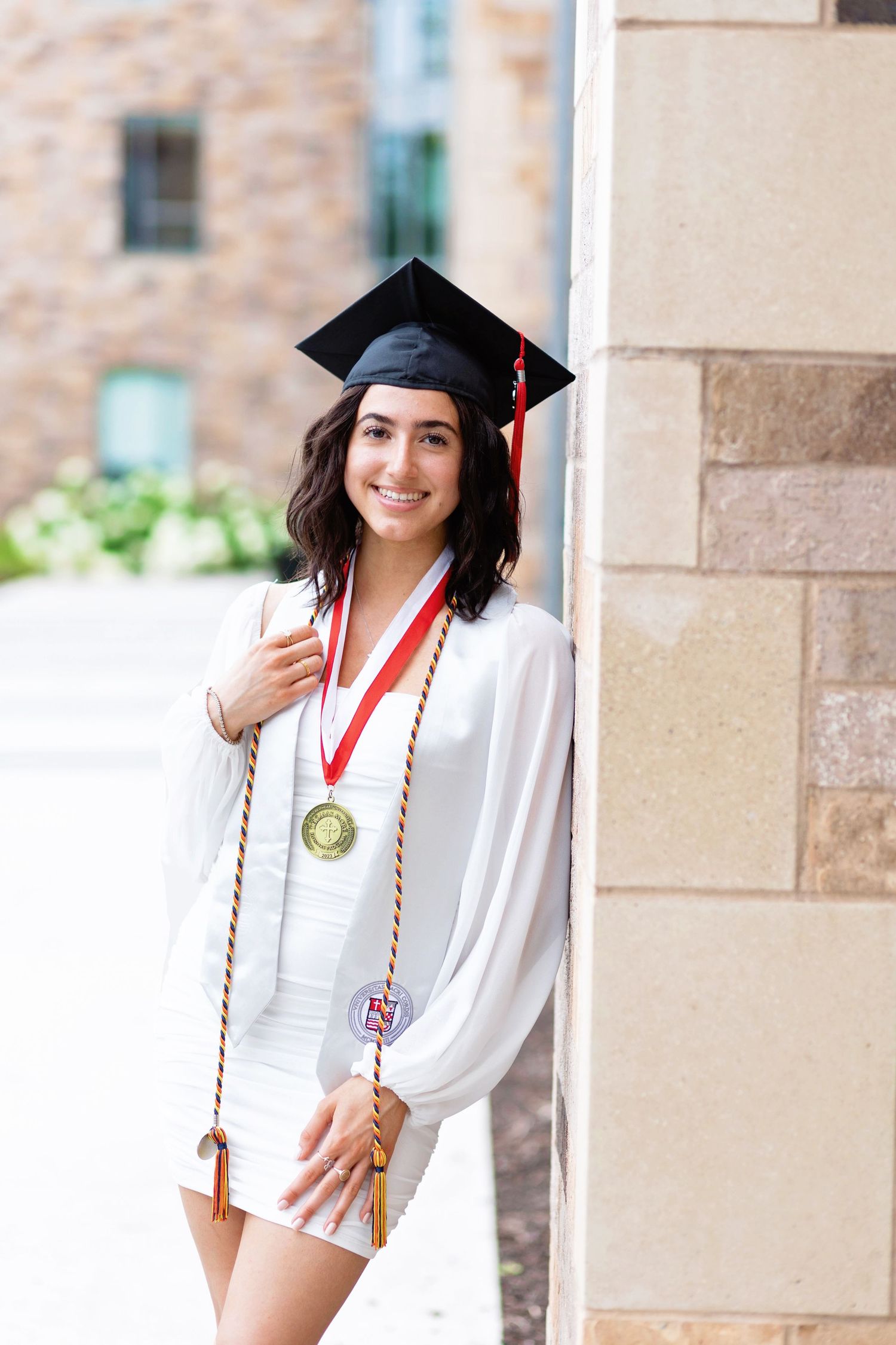 Graduate in white dress and black cap poses against stone wall with honor cords and medallion.
