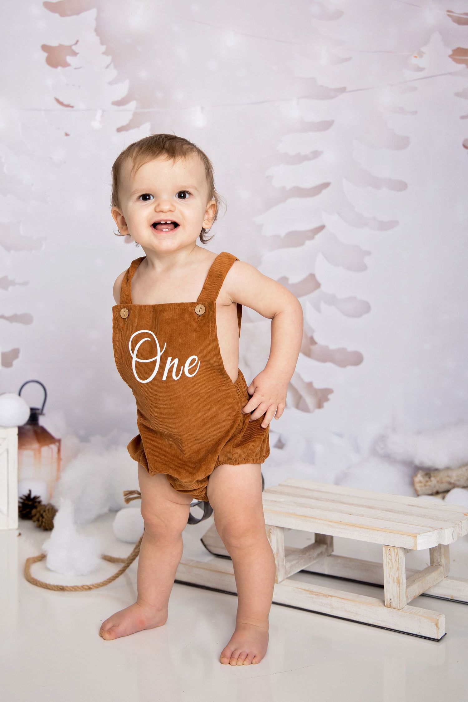 A baby in a brown corduroy romper stands against a white backdrop with watercolor-style wall decorations.