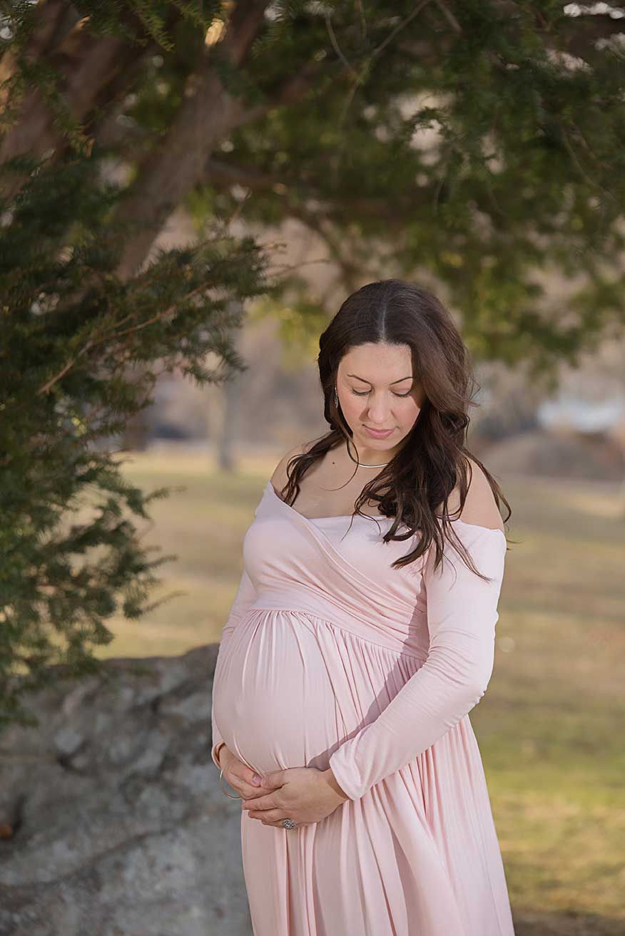 A woman in a soft pink maternity dress poses outdoors under tree branches at golden hour.