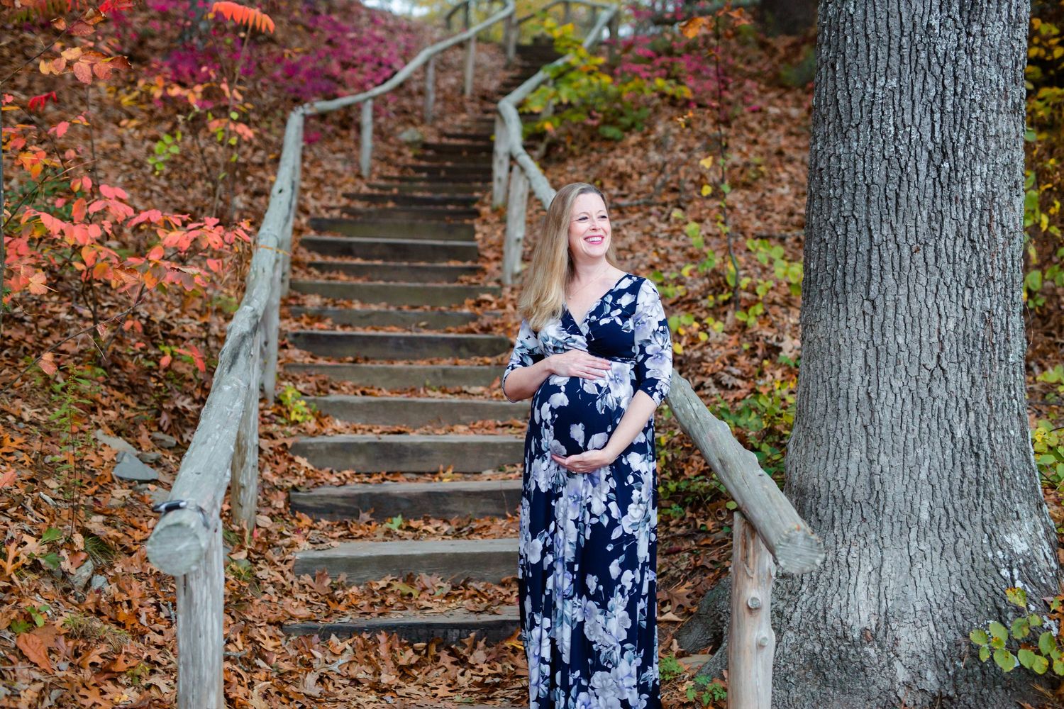 A figure in a navy and white floral dress stands on rustic wooden stairs surrounded by autumn foliage.