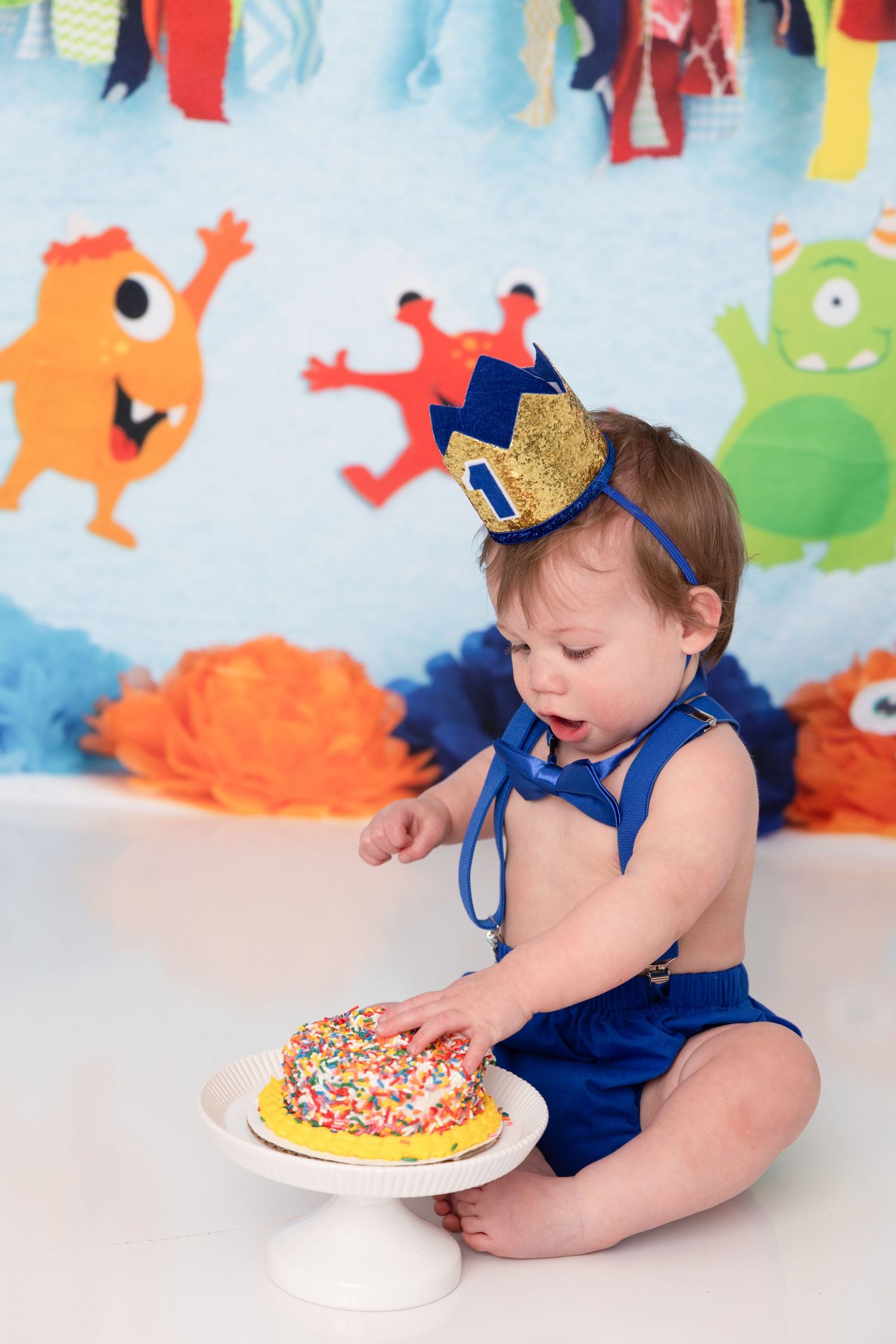 Baby in navy blue outfit reaching for colorful sprinkle-covered cookie or cake during first birthday celebration photoshoot.