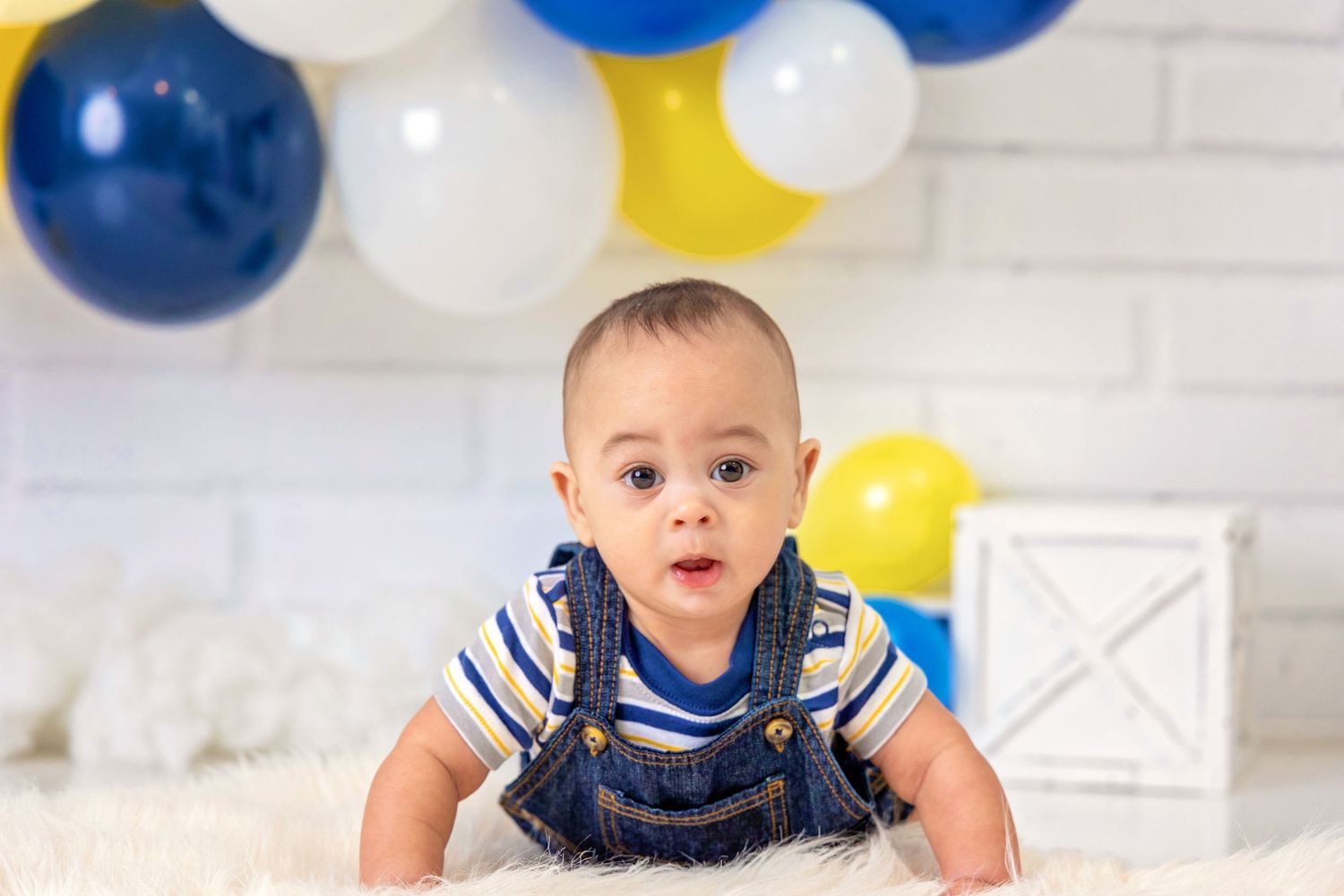 A baby in blue striped clothing crawls on a white floor with blue and yellow balloons in the background.