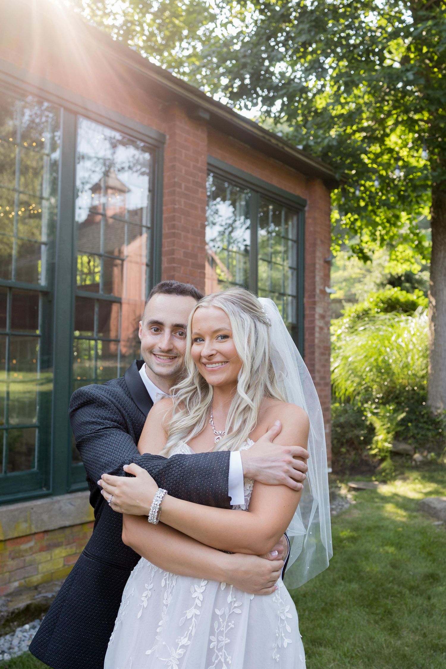 A couple embraces on their wedding day in front of a brick building with green windows and sunlit trees in the background.
