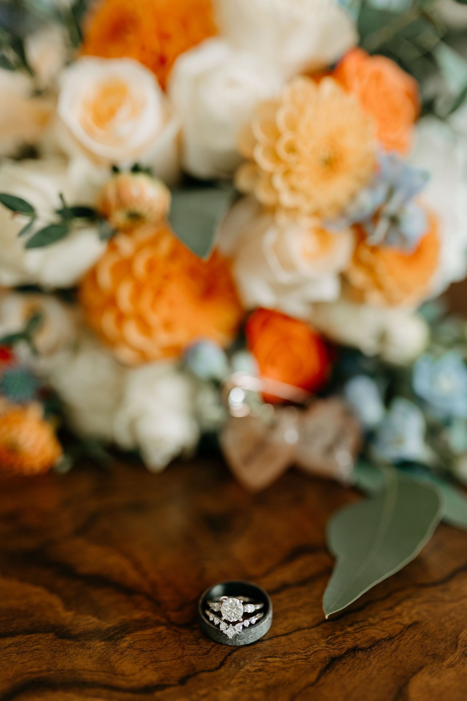Wedding rings displayed with orange and white floral arrangements on wooden surface.