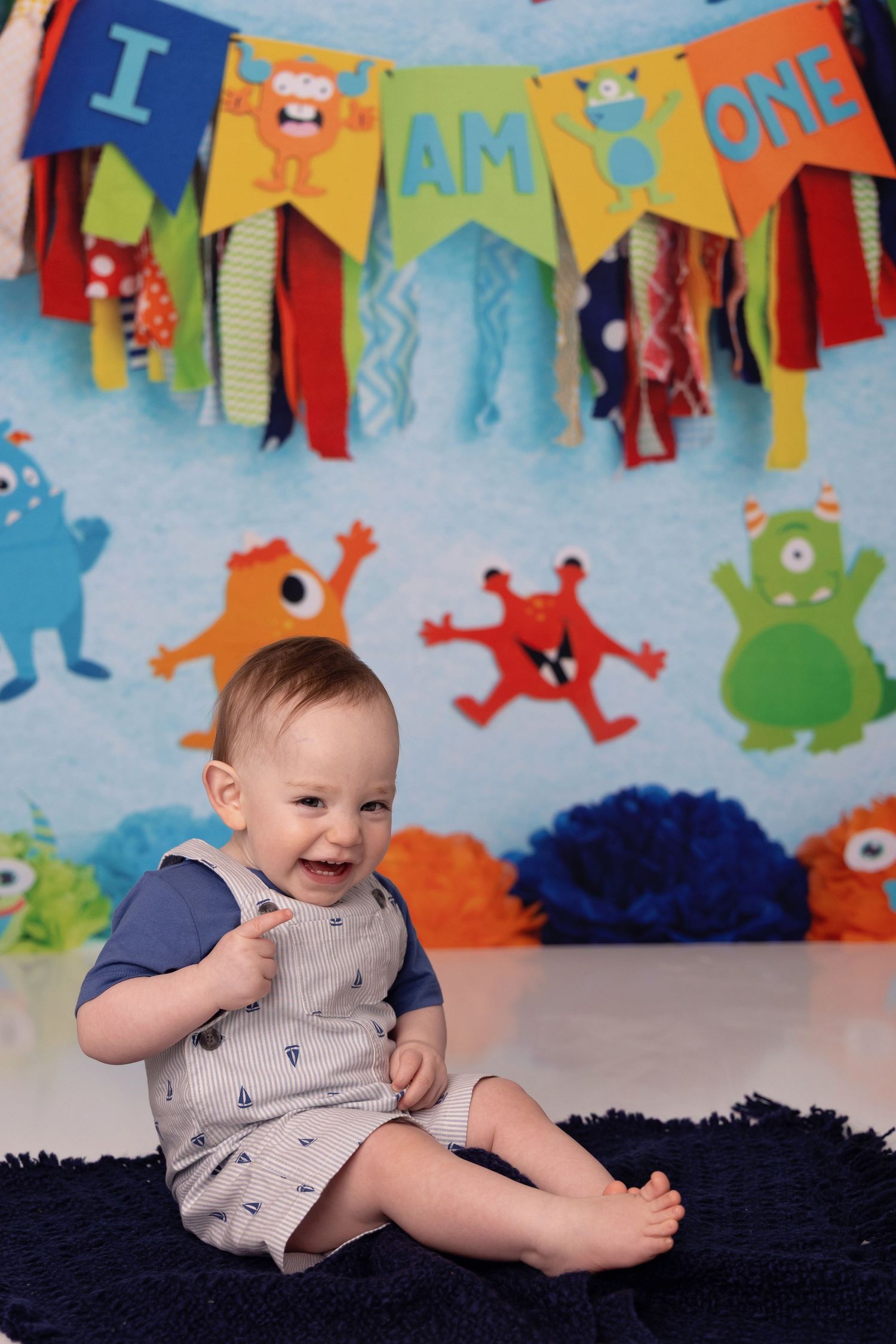 A baby sits on a black rug against a colorful monster-themed party backdrop with festive bunting flags.