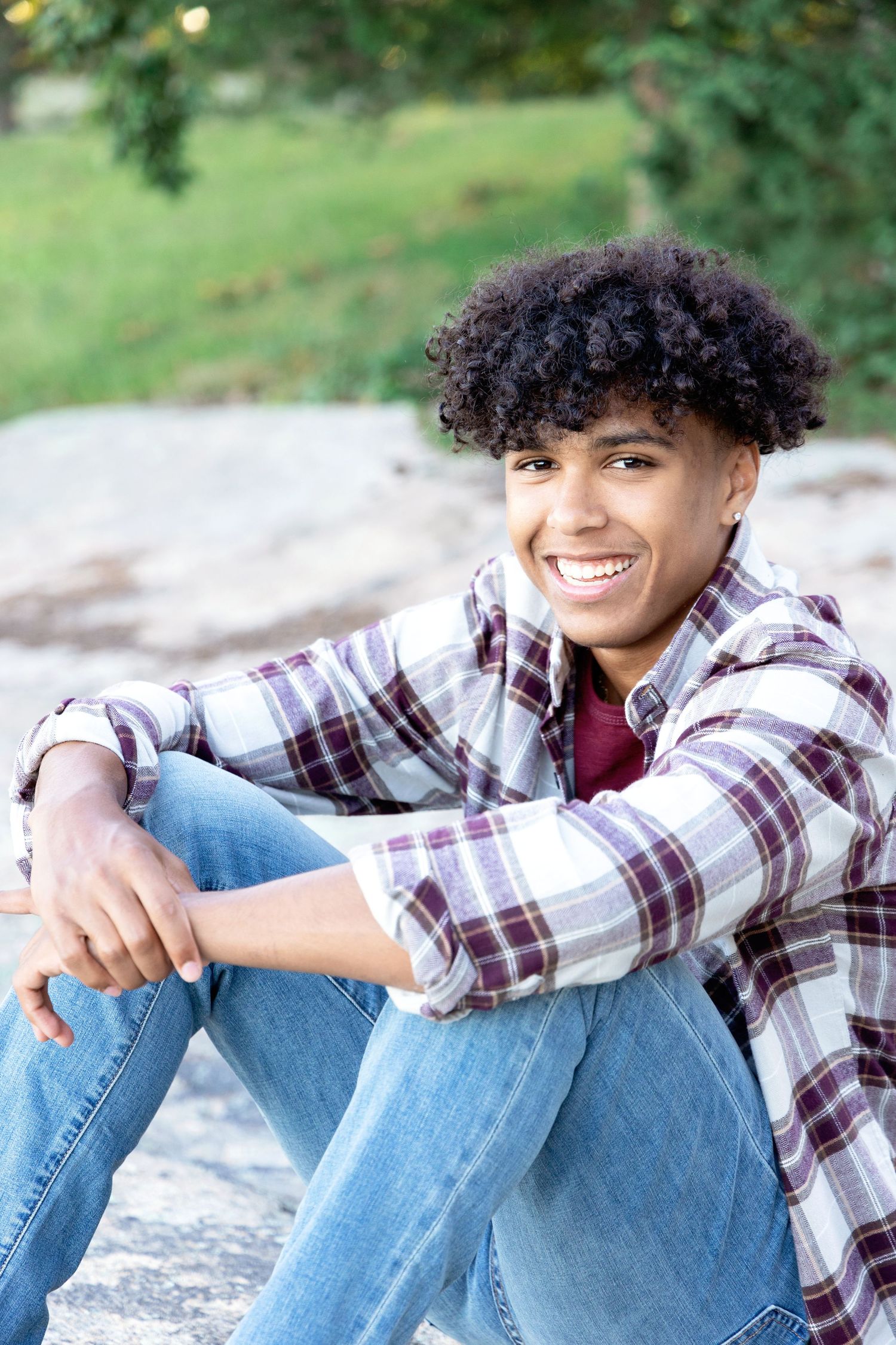 A person in a plaid shirt and jeans sits casually outdoors against a natural backdrop.