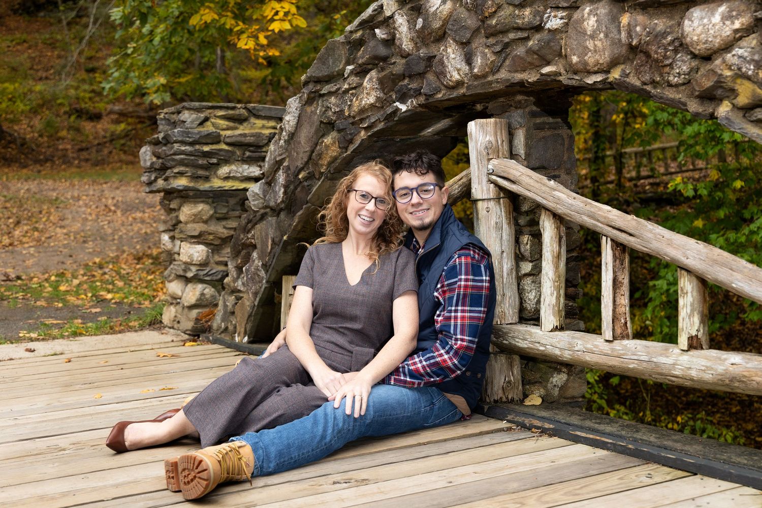 A couple sits together on a rustic wooden bench against a stone wall in an autumn garden setting.