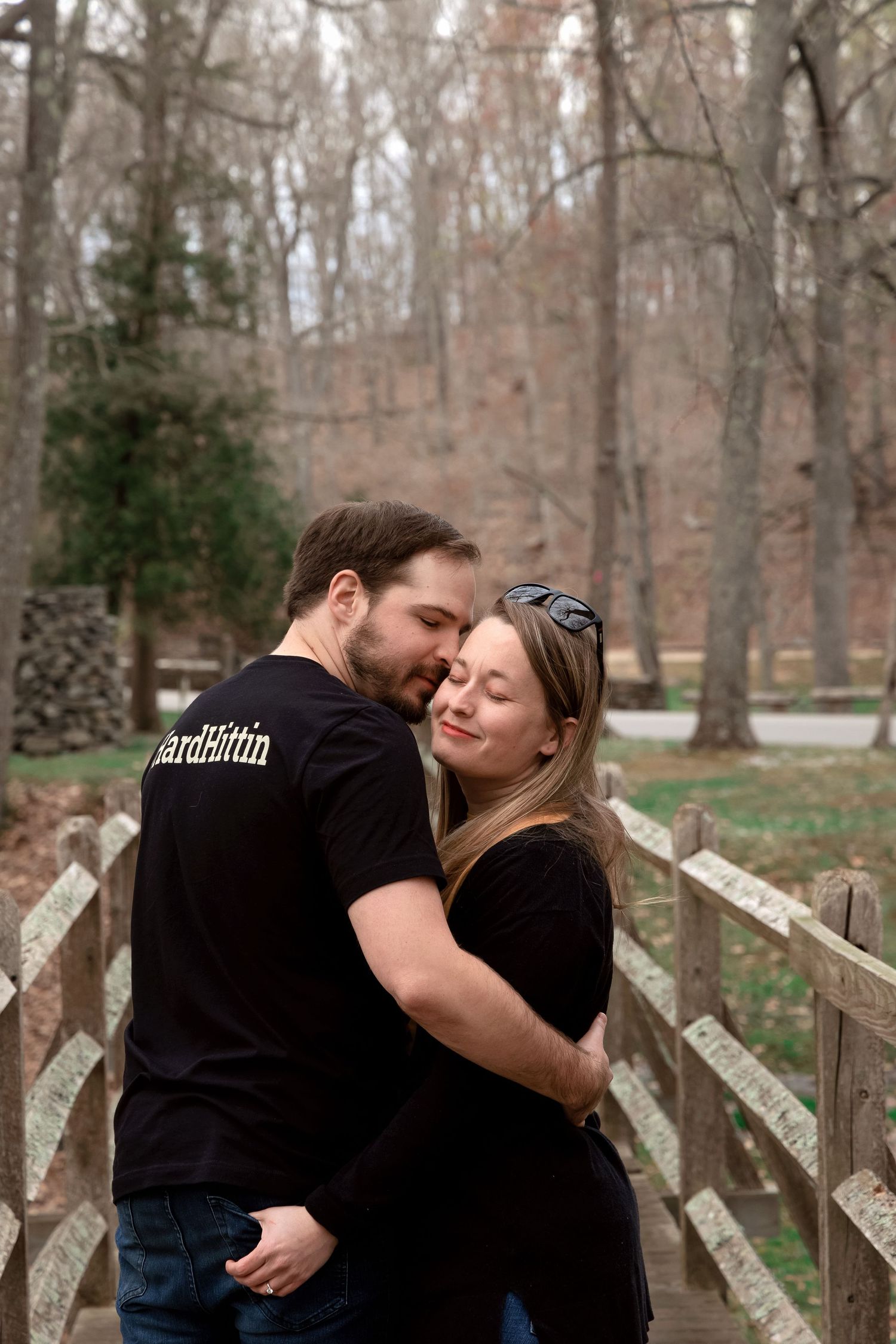 A romantic couple shares a tender kiss while standing near a wooden fence in an autumn forest setting.