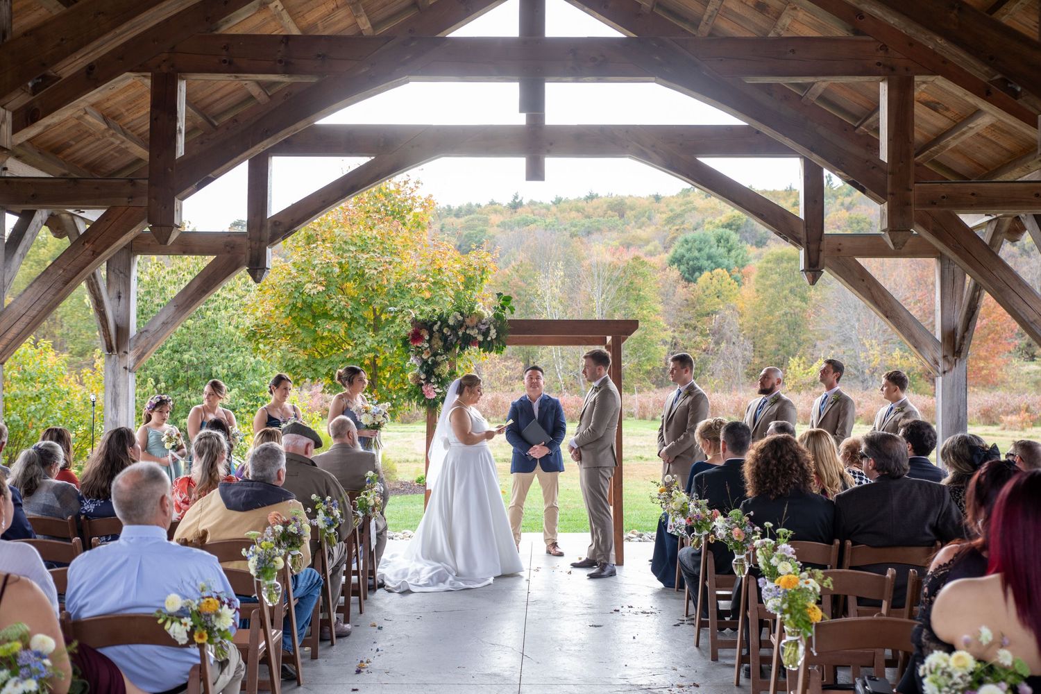 Wedding ceremony taking place under a rustic wooden pavilion surrounded by autumn foliage and guests.