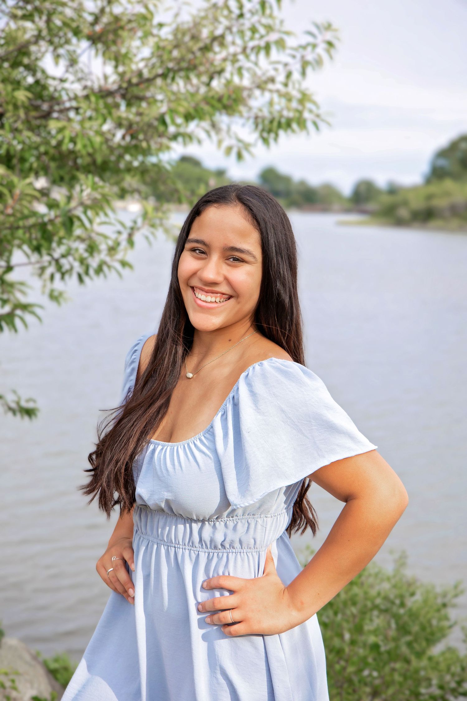 Young person in light blue summer dress poses outdoors by a lake with trees in the background.