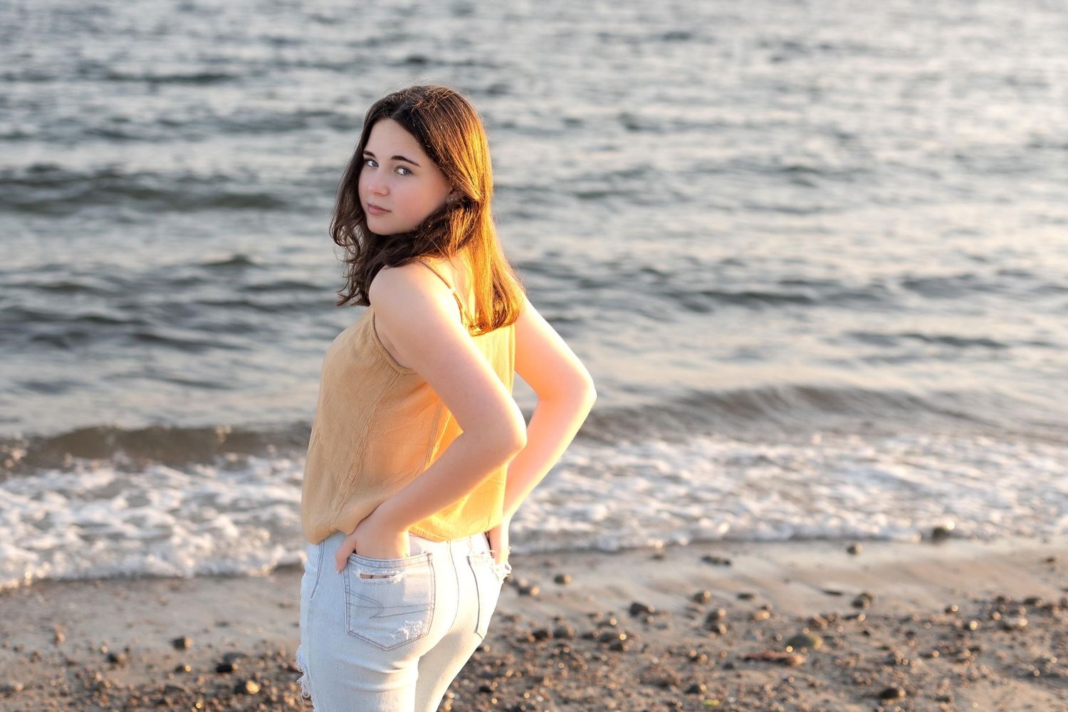 Person in a white top stands on a rocky beach at sunset as waves roll onto the shore.