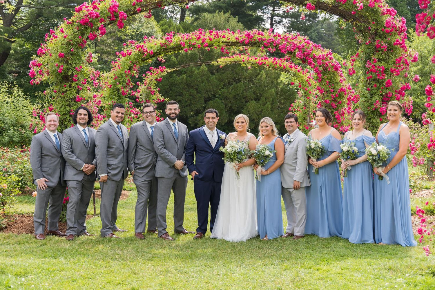 Wedding party poses together in front of vibrant pink flowers and green foliage in an outdoor garden setting.