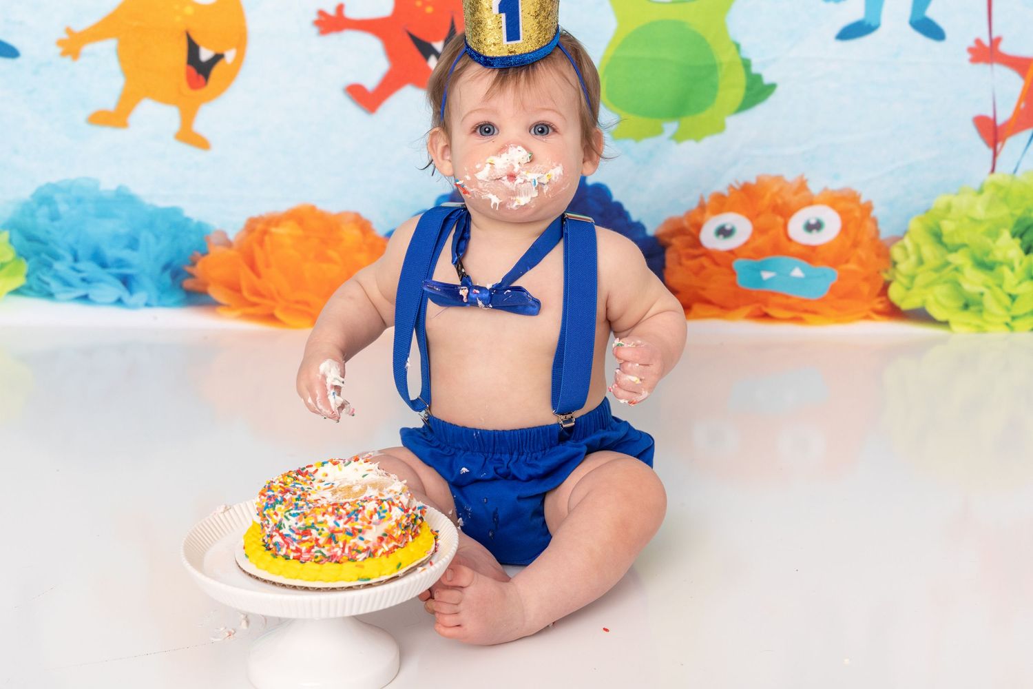 Baby in navy blue outfit sits on floor with colorful cake smash setup and festive decorations for first birthday celebration.