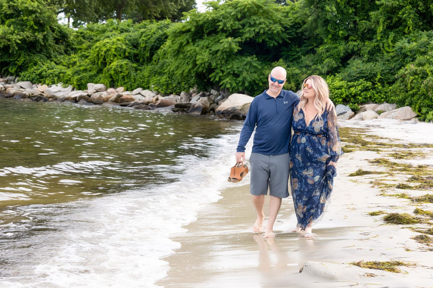 Two people wade through shallow river water surrounded by lush green trees and rocks along the riverbank.