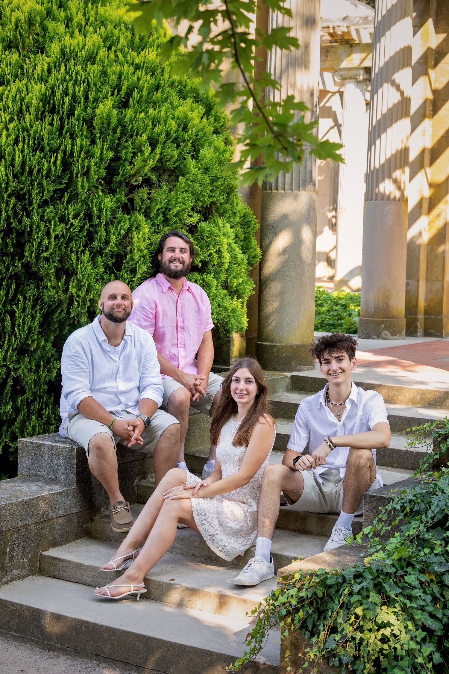 People sit casually on stone steps surrounded by lush green bushes in a garden setting.