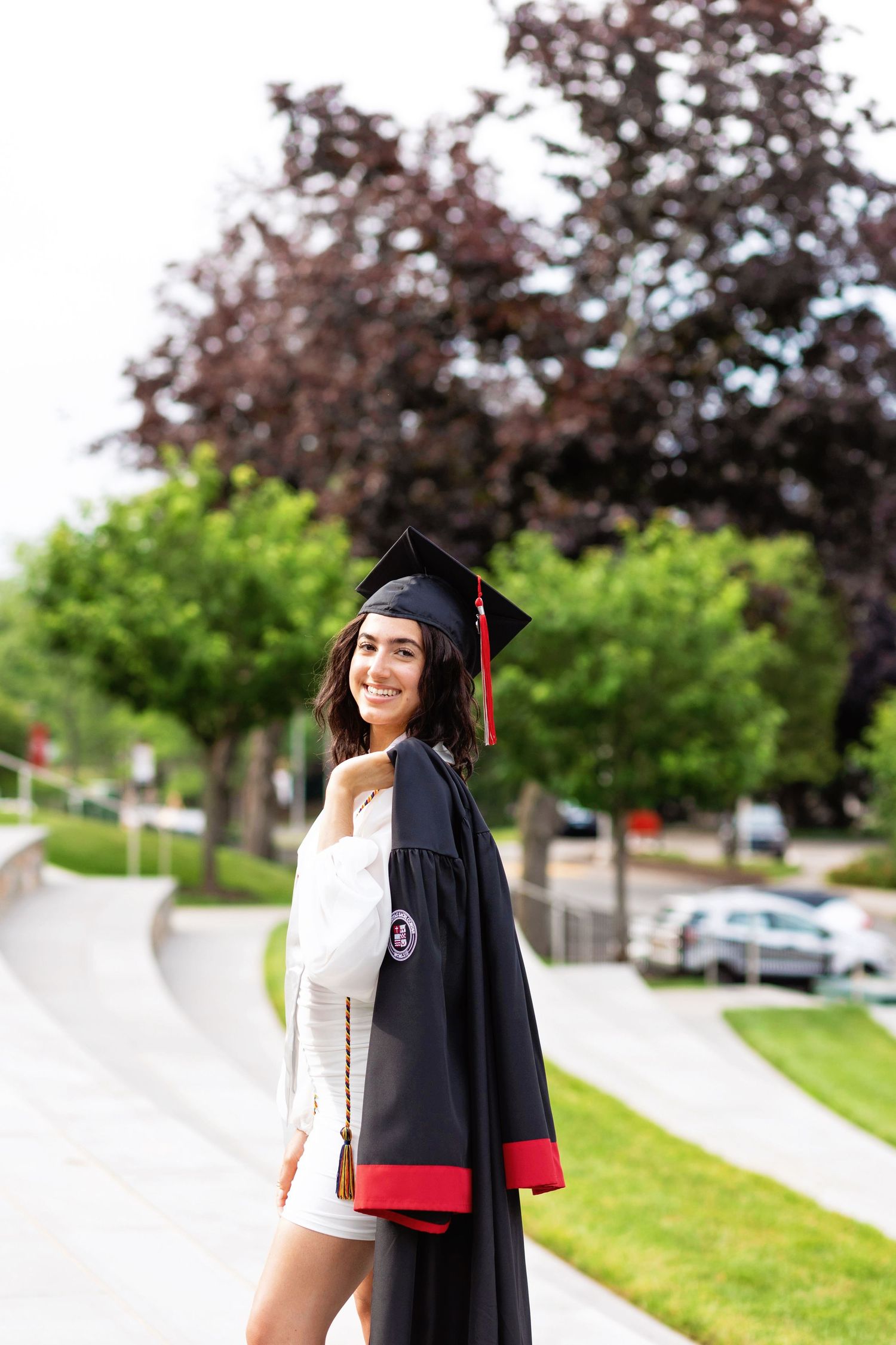 Graduate in cap and gown smiles while standing on campus sidewalk with trees in background.
