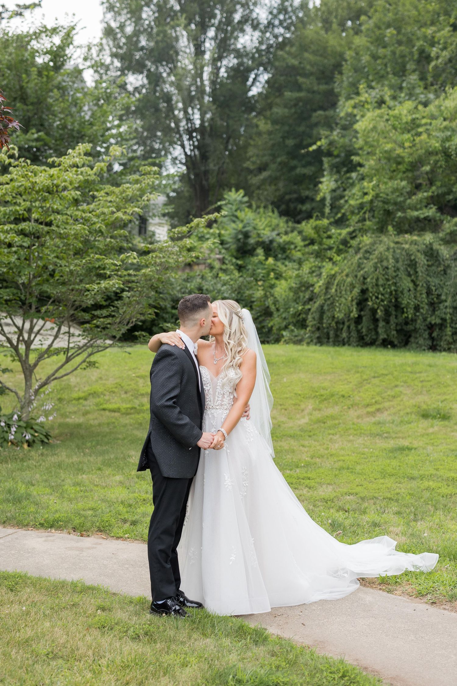 A couple shares a romantic moment on a winding path surrounded by lush green trees and grass in a garden setting.