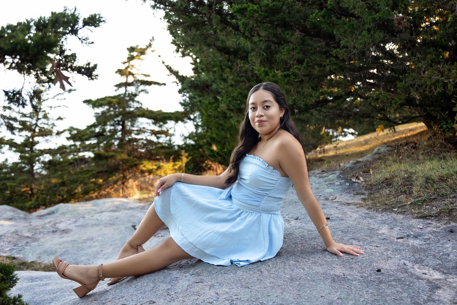 A person in a flowing light blue dress poses gracefully on a rocky cliff during golden hour.