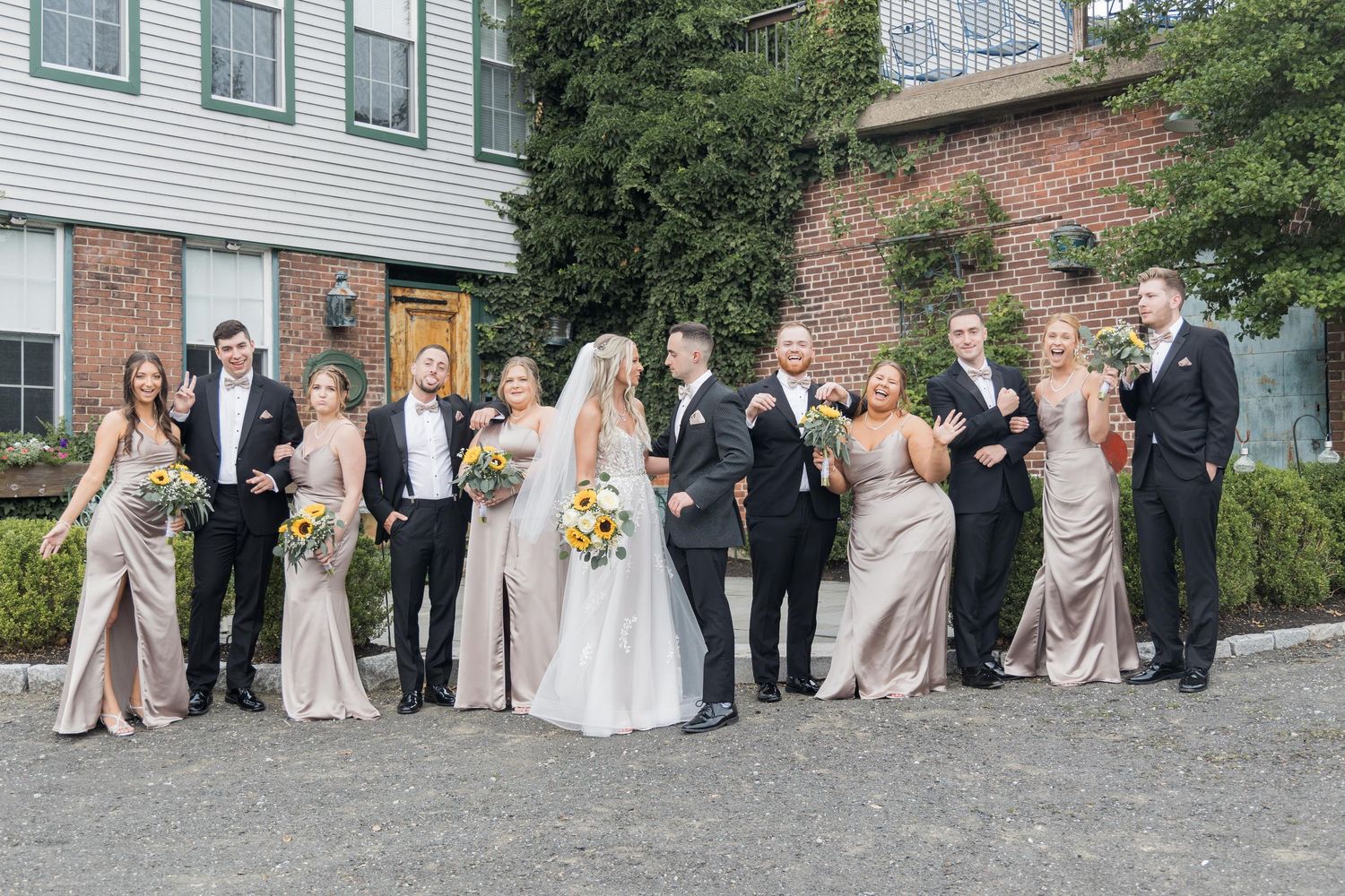 Wedding party lined up outdoors with bridesmaids in beige dresses and groomsmen in dark suits.