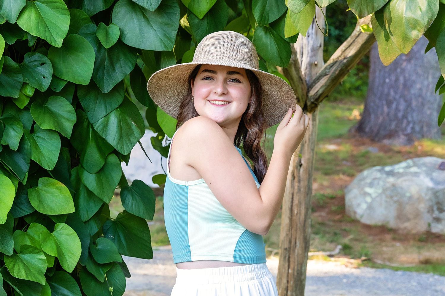 A person wearing a sun hat smiles while standing among lush green foliage in a garden setting.