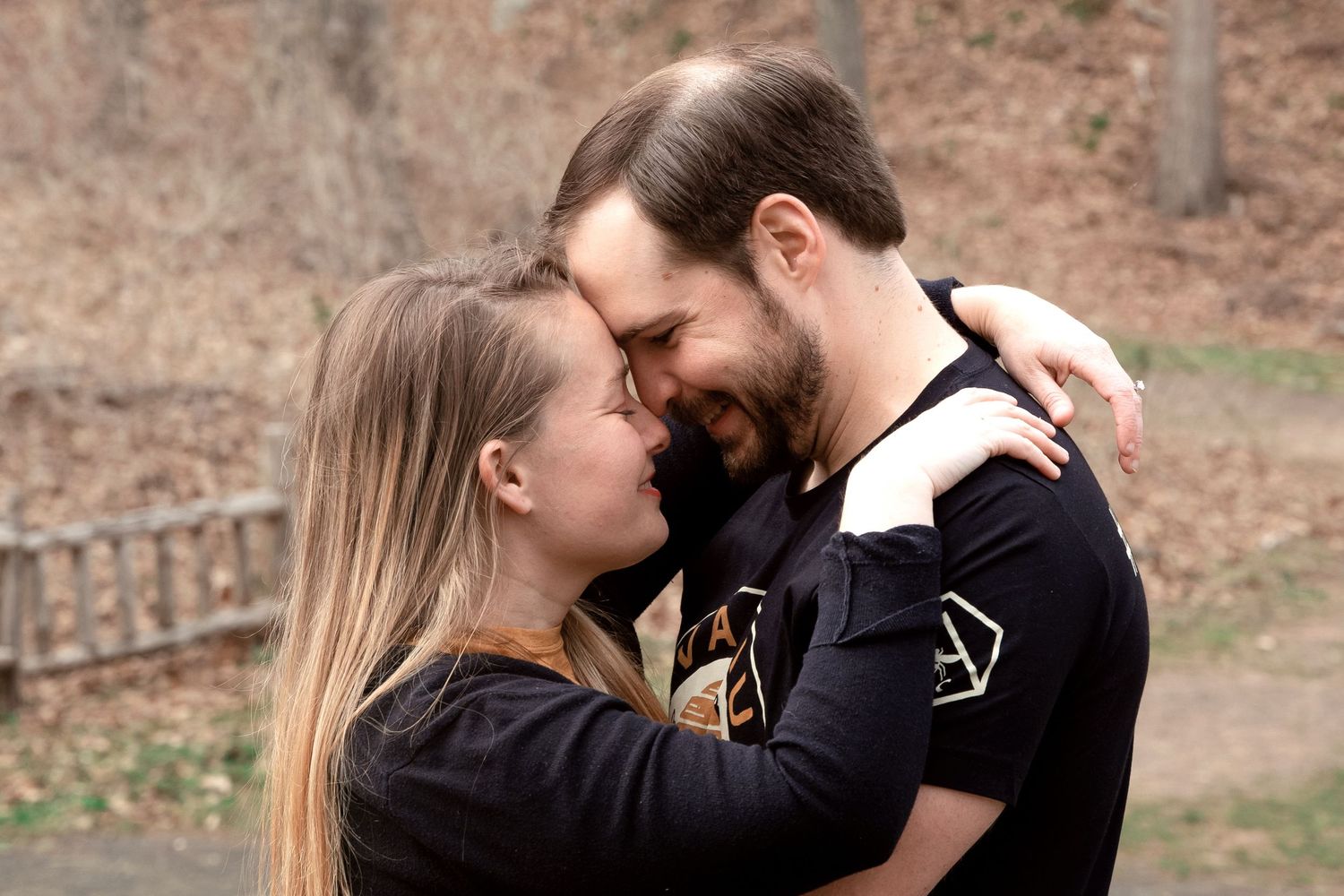 A couple embraces tenderly outdoors in autumn, wearing black clothing and sharing an intimate moment together.