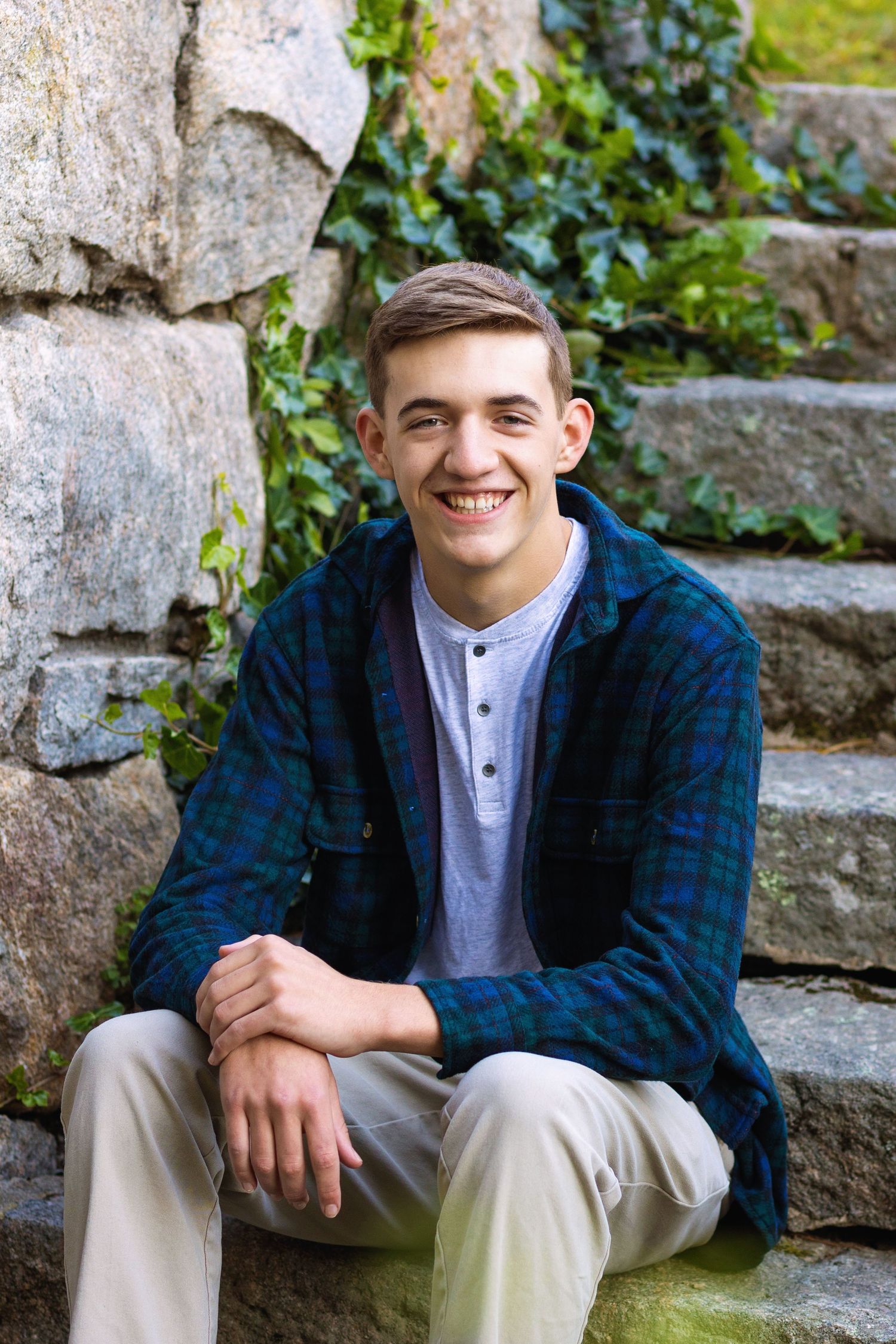 A person in a blue plaid shirt and white henley sits casually on stone steps outdoors with greenery in the background.