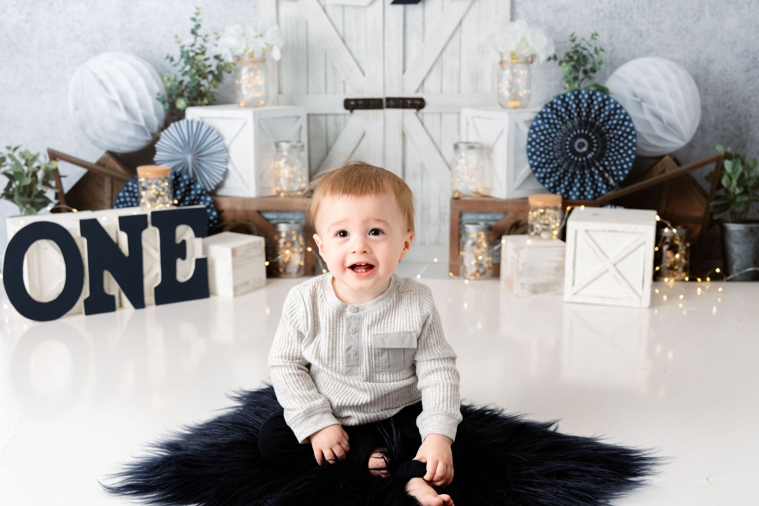 A baby in gray clothing sits on a white backdrop with blue and silver party decorations for a first birthday celebration.