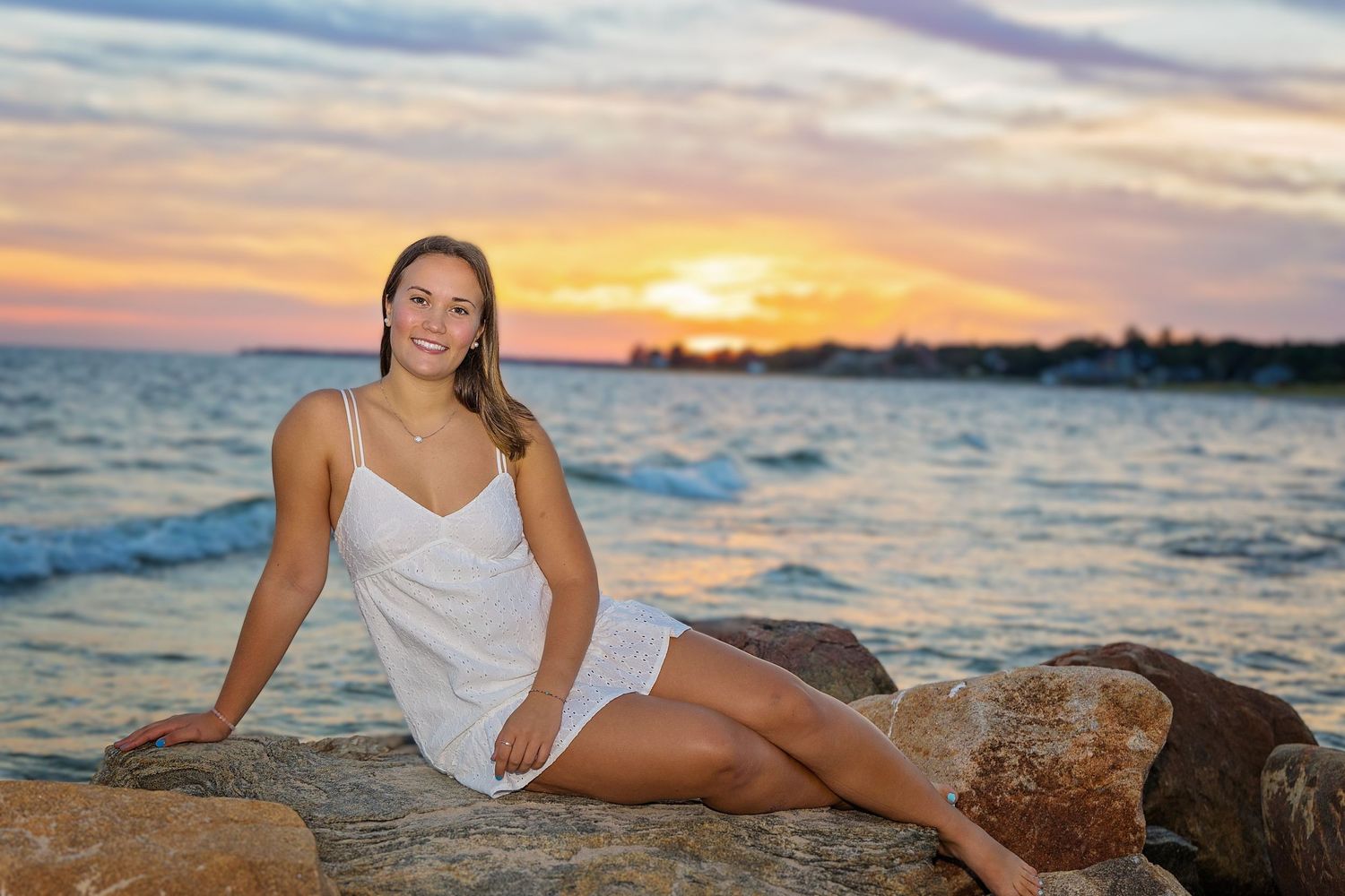 A person in a white dress poses on coastal rocks at sunset as waves crash against the shore.