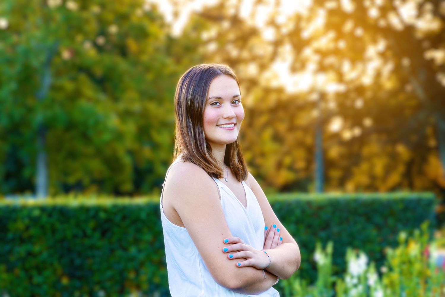A person in a white sleeveless top poses with arms crossed in a garden during golden hour sunset.