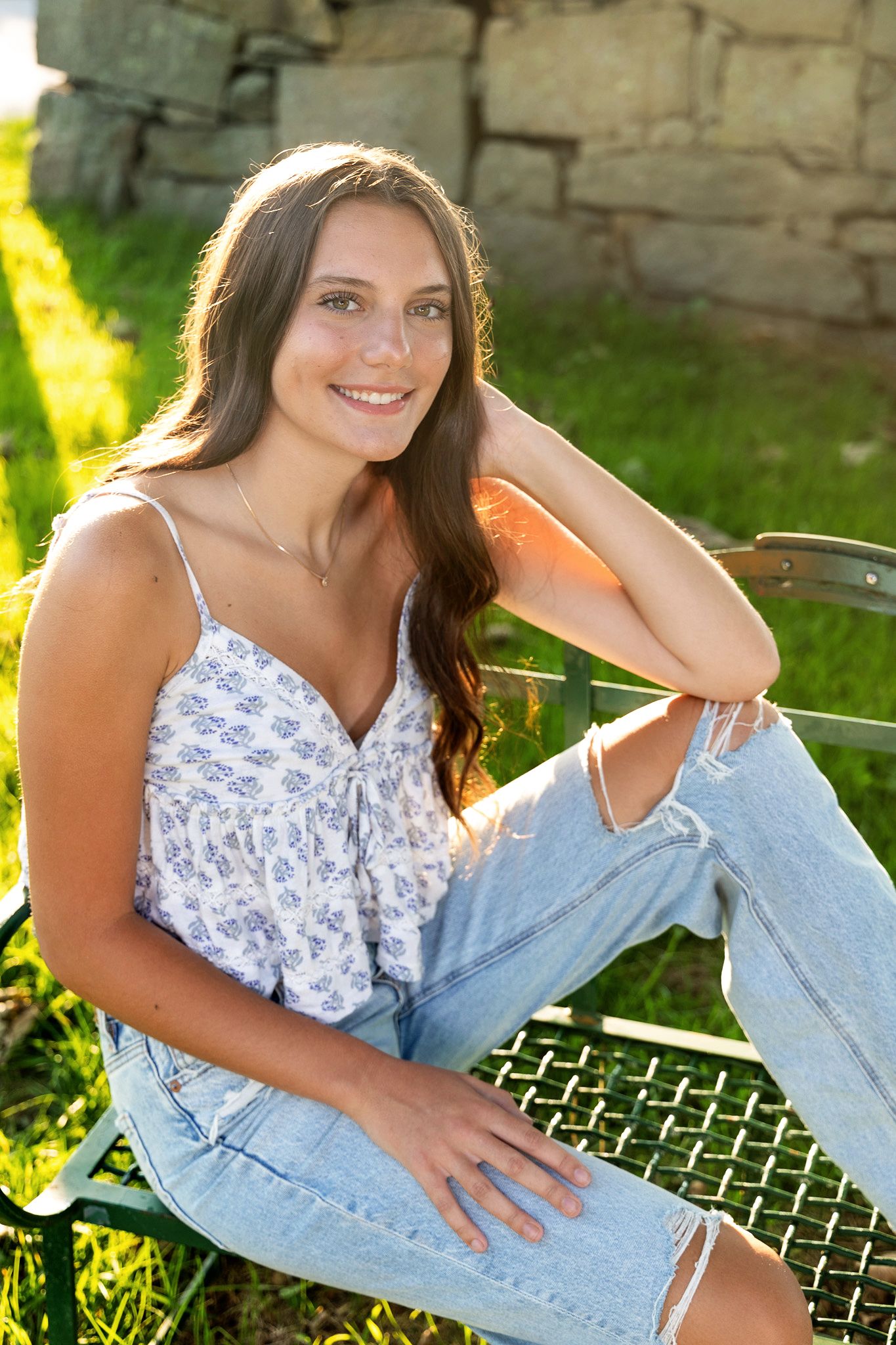 Casual outdoor photo session on a stone bench with blue jeans and a floral top against natural lighting.