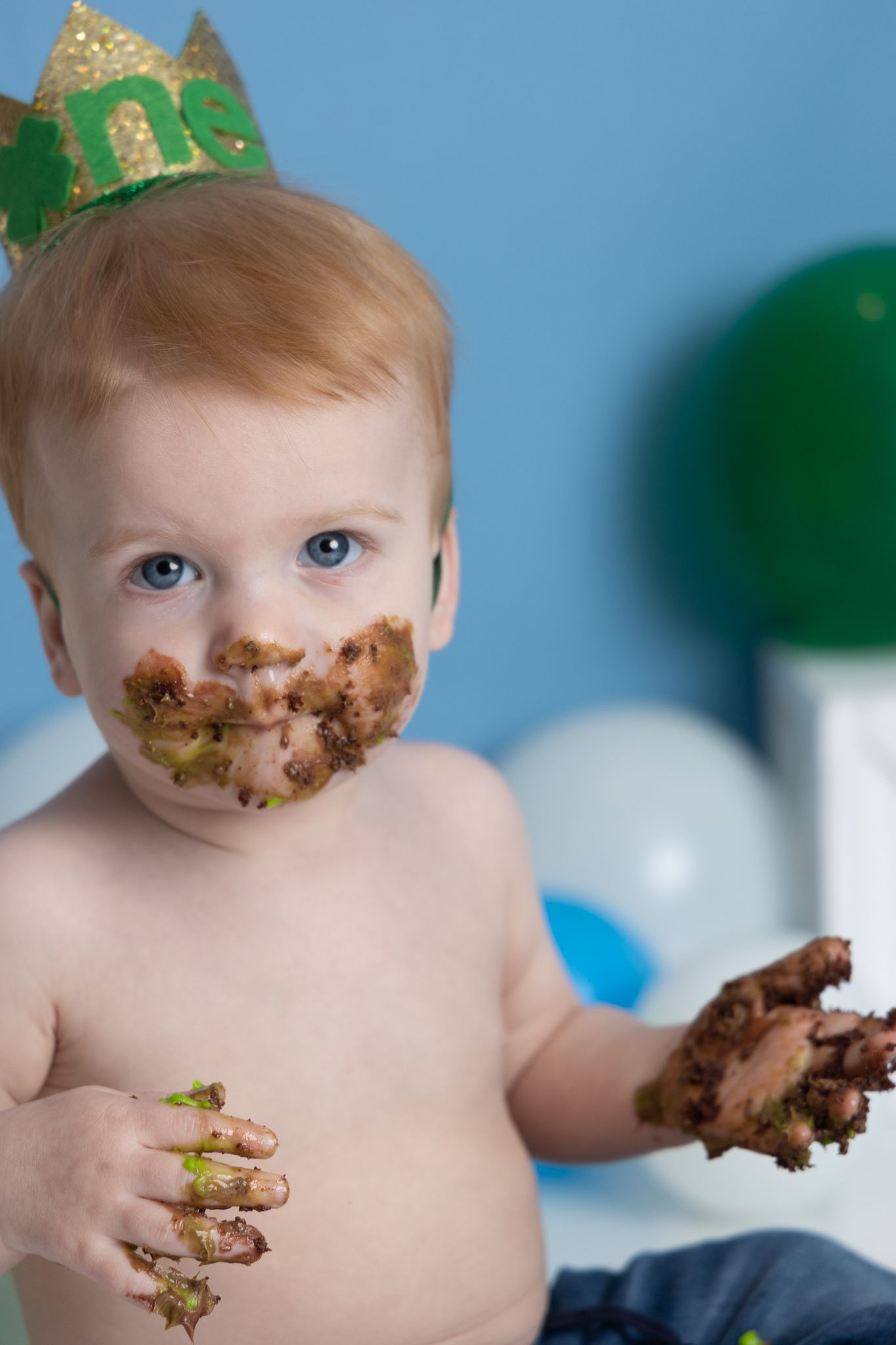 A small child wearing a gold crown enjoys a messy chocolate cake during a birthday celebration with balloons in the background.