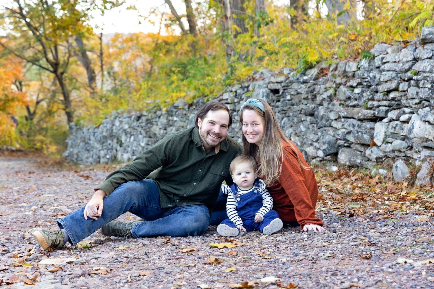 A family sits together on fallen autumn leaves near a stone wall in a park during golden hour.