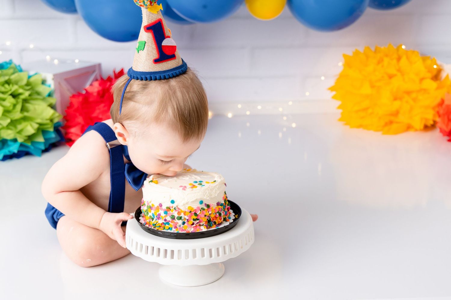 Baby in a party hat leans over to taste birthday cake while surrounded by blue balloons and colorful decorations.