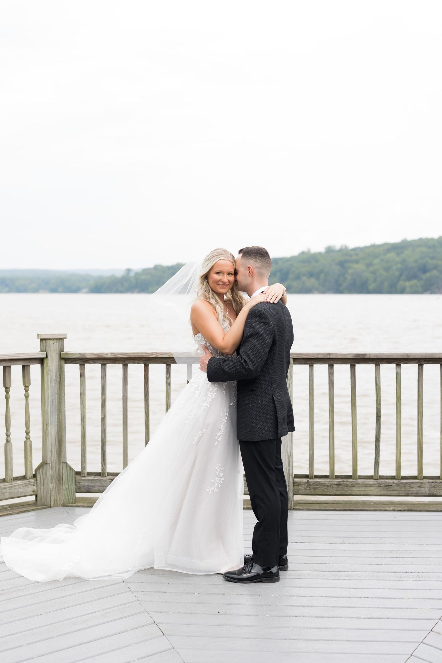 A romantic wedding couple shares intimate moments on a lakeside deck with scenic water views in the background.