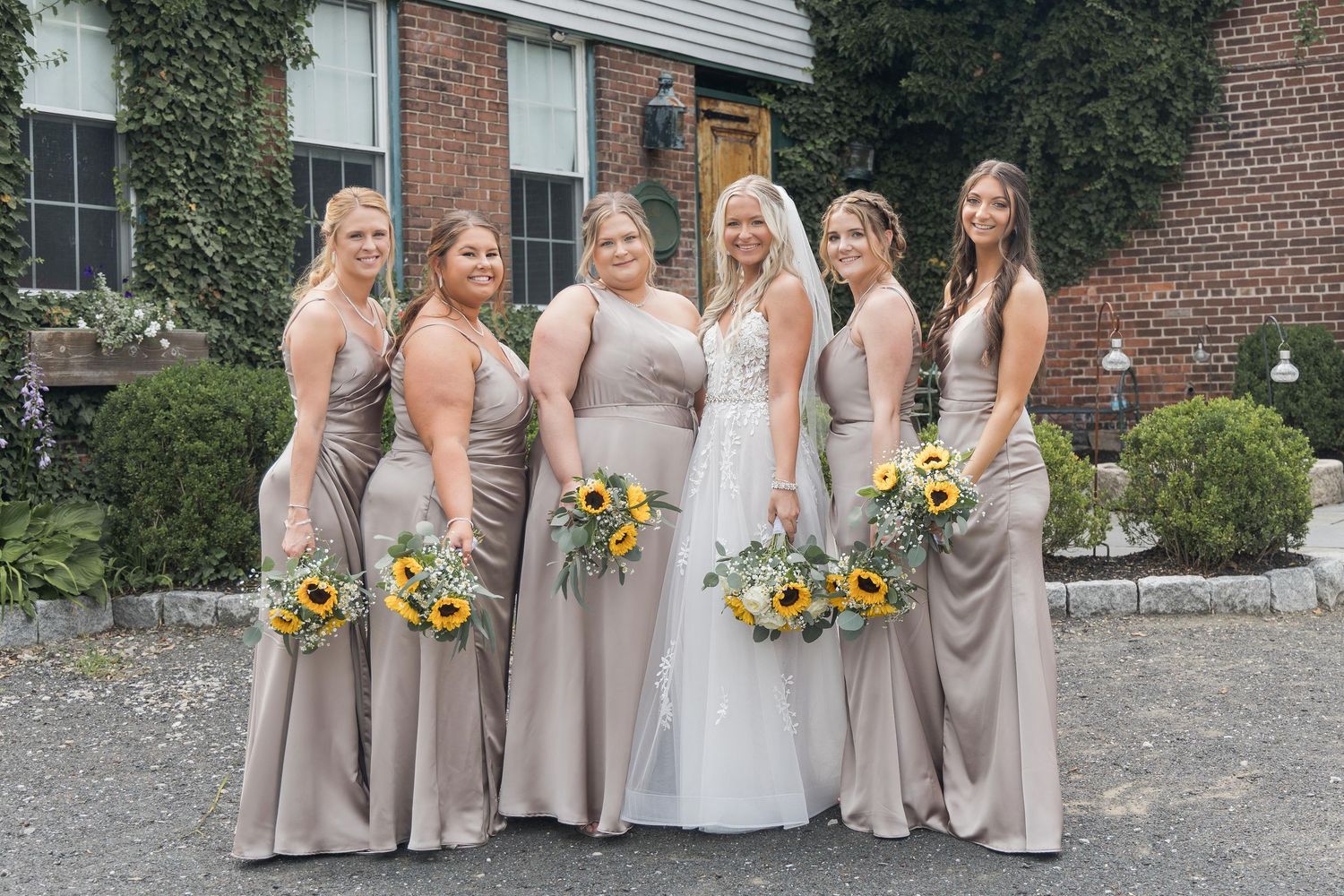 A wedding party in neutral beige dresses poses together holding yellow sunflower bouquets outdoors by a brick building.
