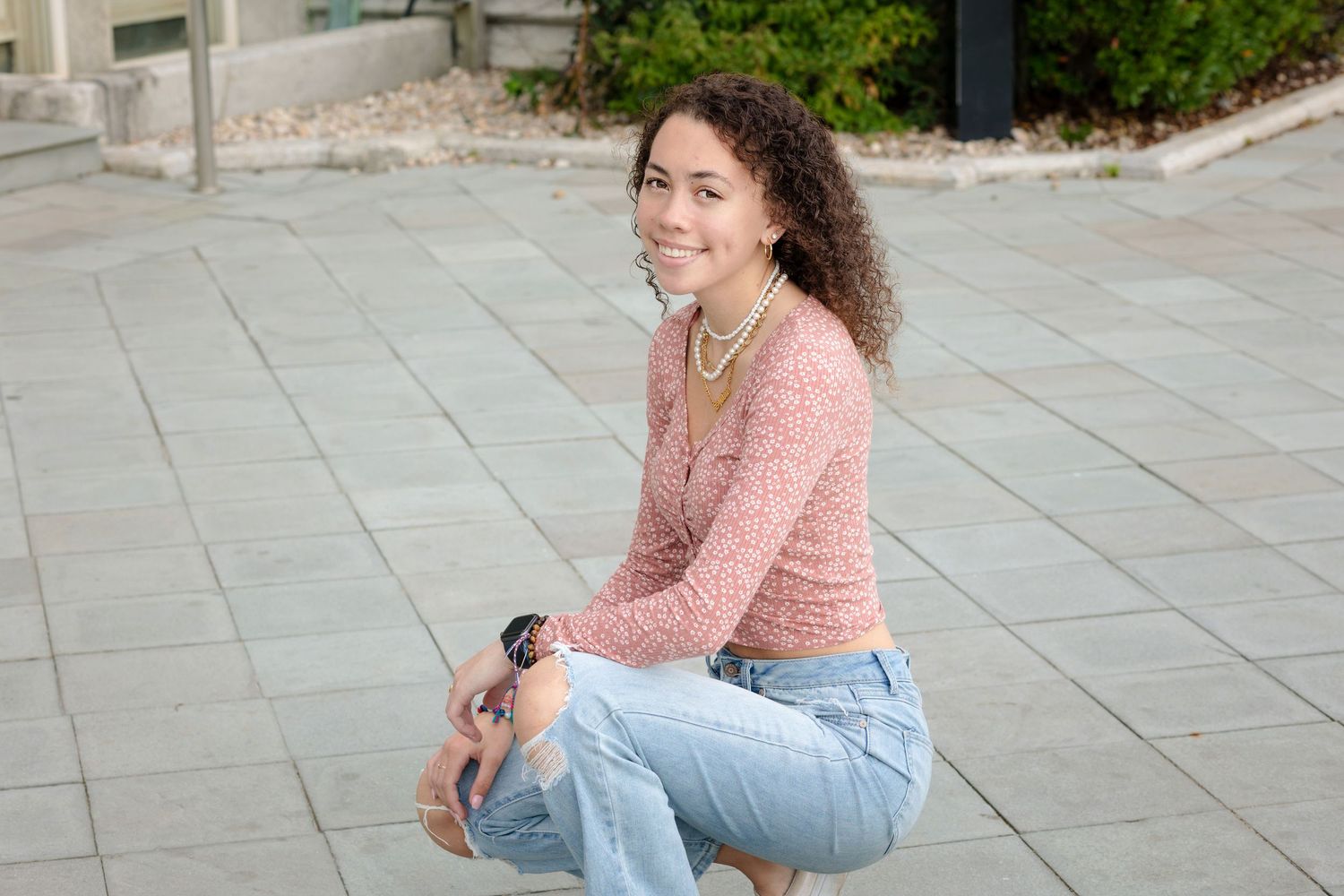 A person in a pink sweater and light jeans sits casually on stone pavement outdoors.