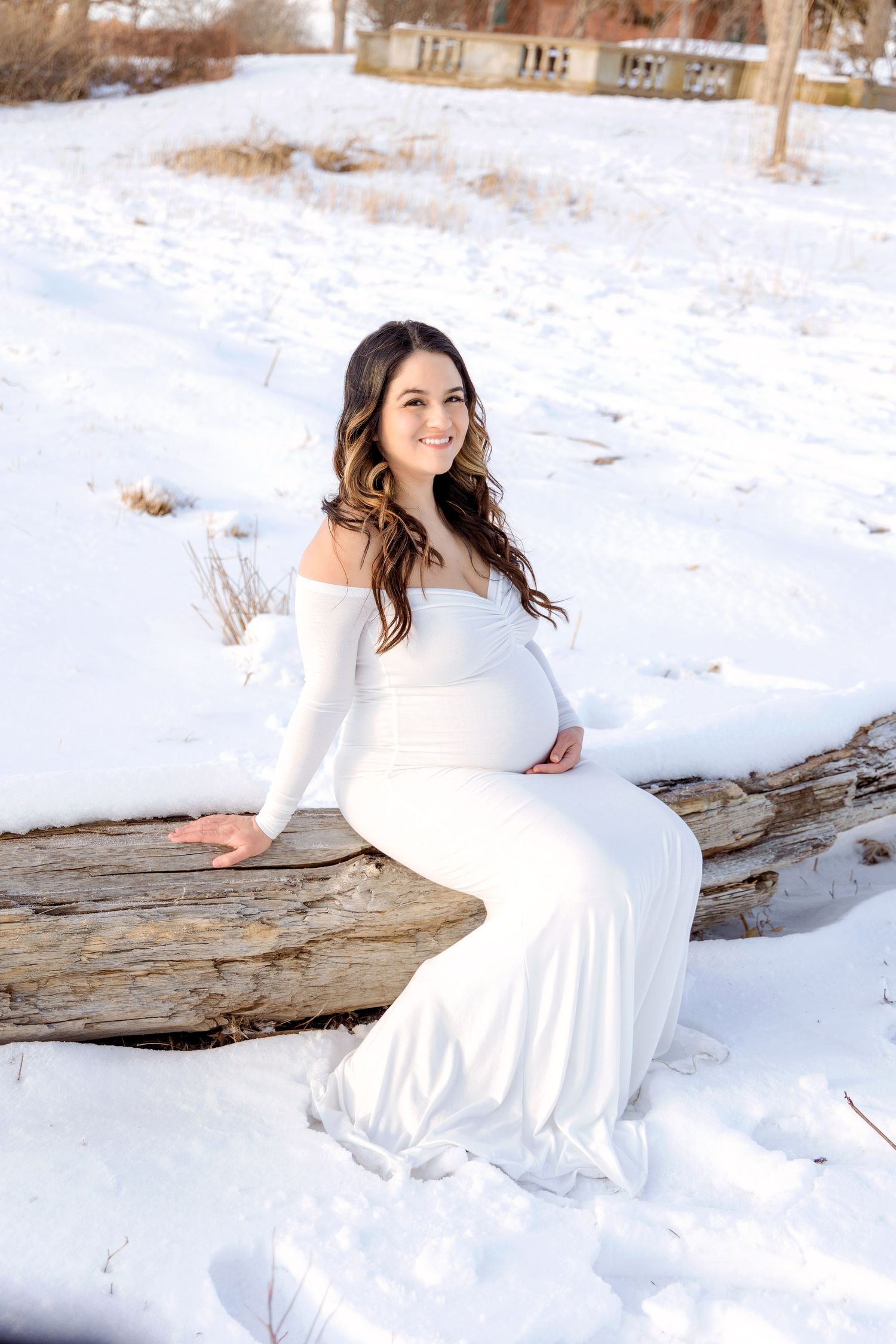 A pregnant woman in a white dress poses on a fallen log in a snowy winter landscape.