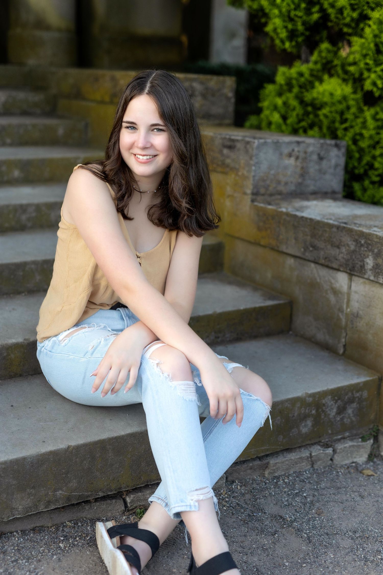 A person in light blue jeans sits casually on stone steps outdoors with greenery visible in the background.